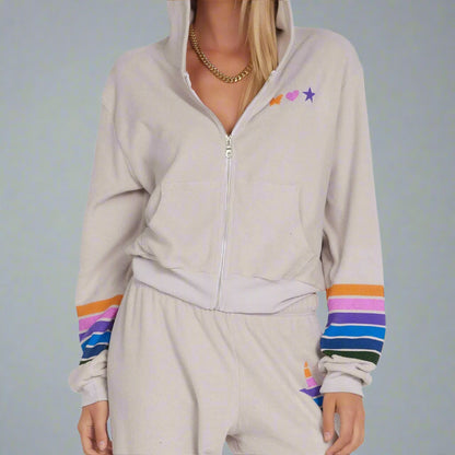 A female model wearing a beige hooded sweatshirt with multicolored stripes and stars on the sleeves.